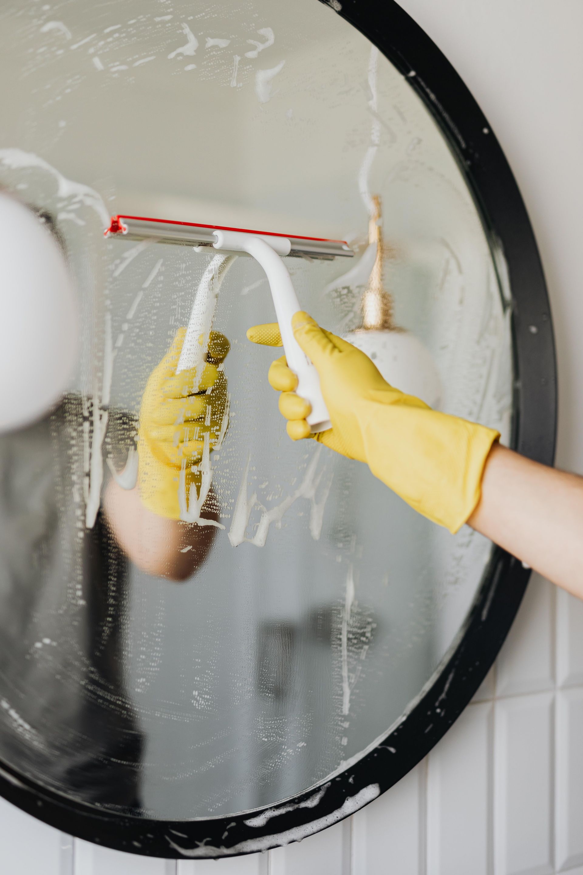 Faceless housekeeper in yellow rubber gloves cleaning round mirror from foam by squeegee in bathroom