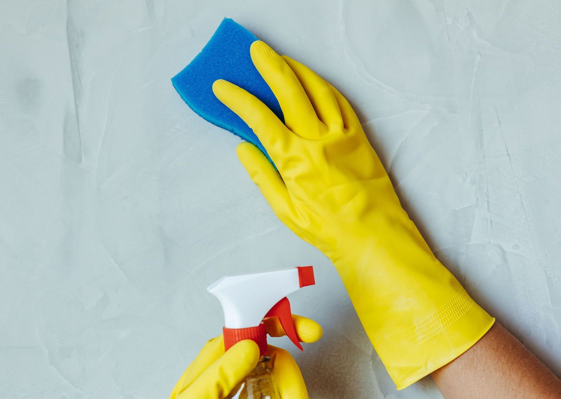 Hands in yellow gloves cleaning wall with sponge and spray bottle.