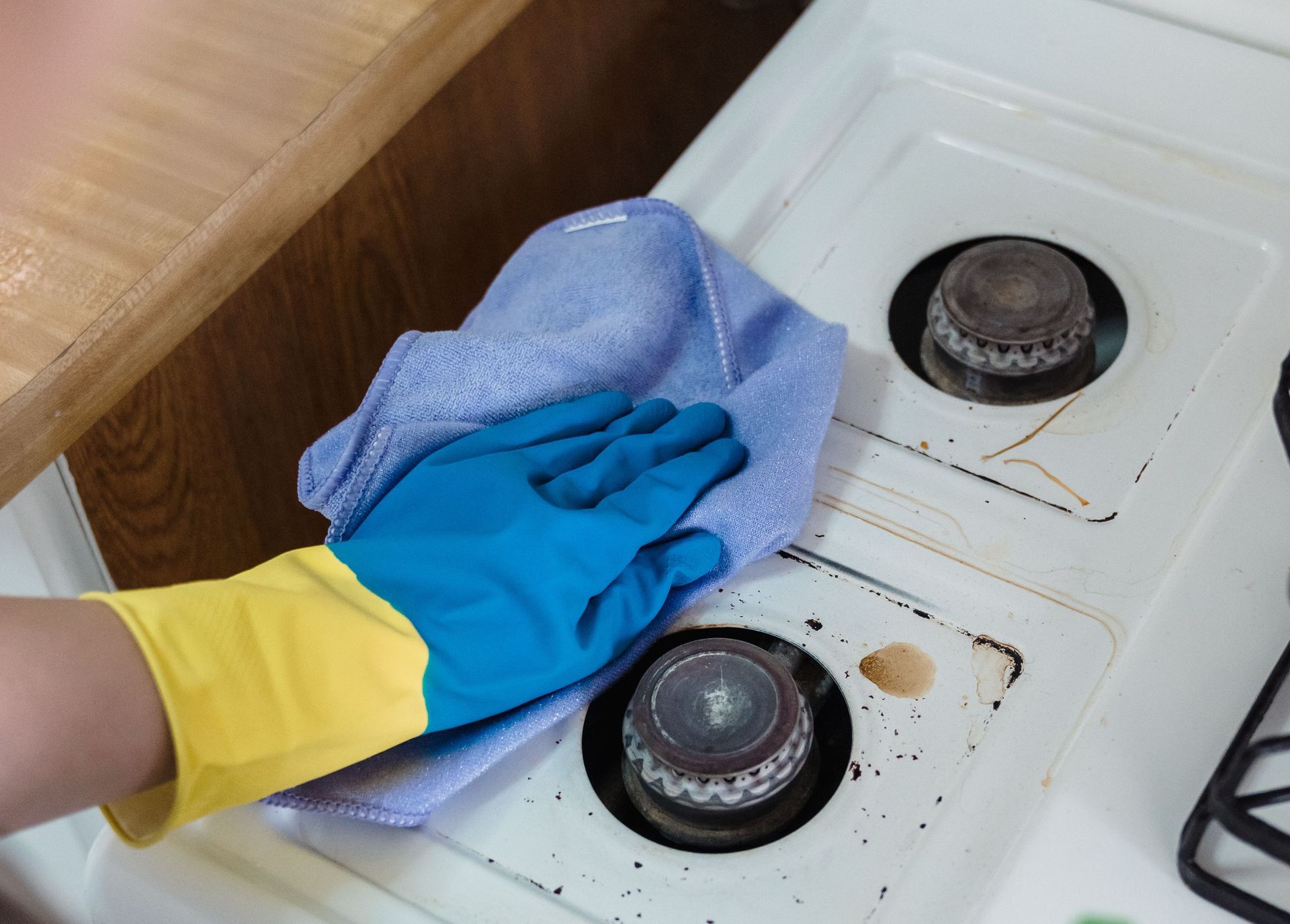 A person wearing rubber gloves cleaning a dirty gas stove with a cloth and spray bottle.